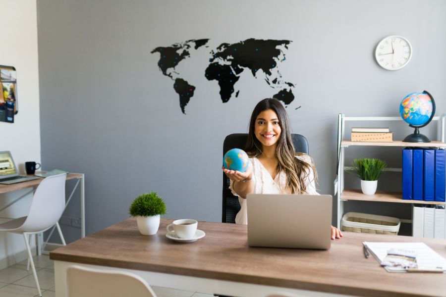 Conseillère en voyage organisé souriante tenant un globe terrestre dans son bureau avec une carte du monde en arrière-plan.
